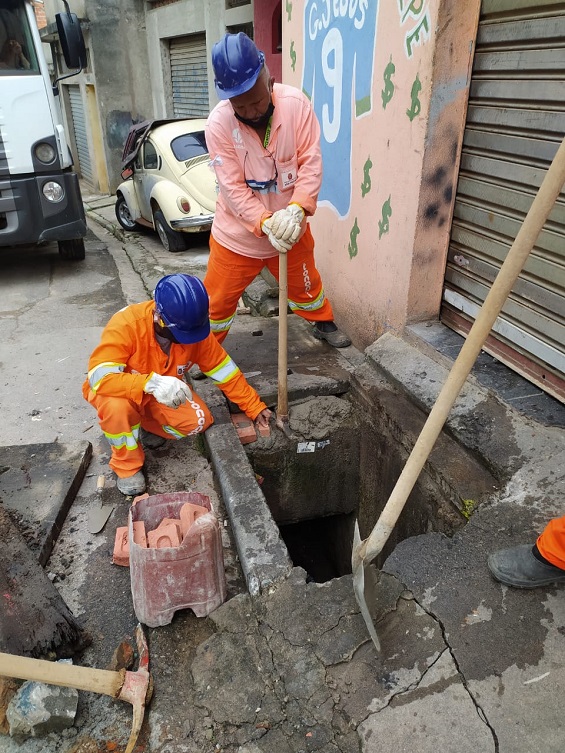 Dois funcionários com macacão laranja trabalham na remoção de uma tampa de concreto e na limpeza de uma boca de lobo.